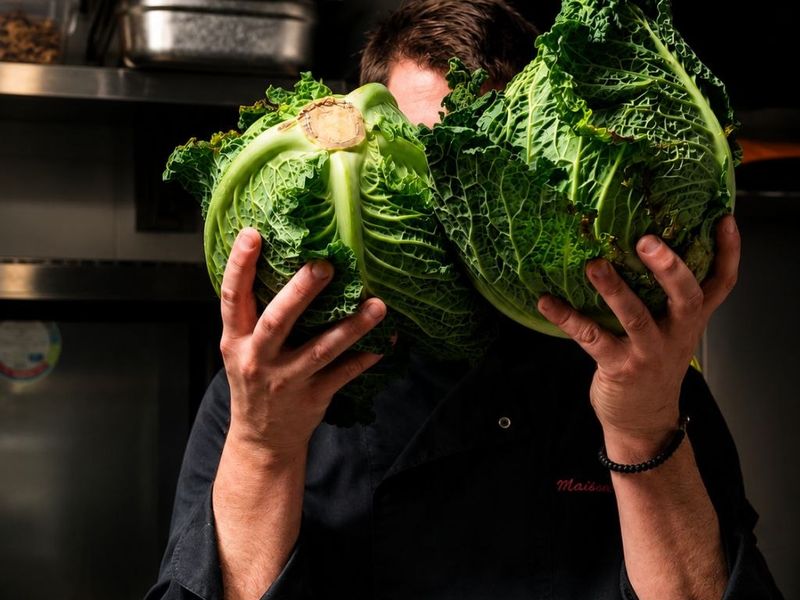 Le chef de Maison Chapron dans sa cuisine, tenant deux choux verts frais devant lui